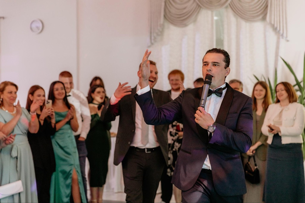 Hochzeitsmoderator moderiert mit Mikrofon die Feier, umgeben von applaudierenden G&auml;sten